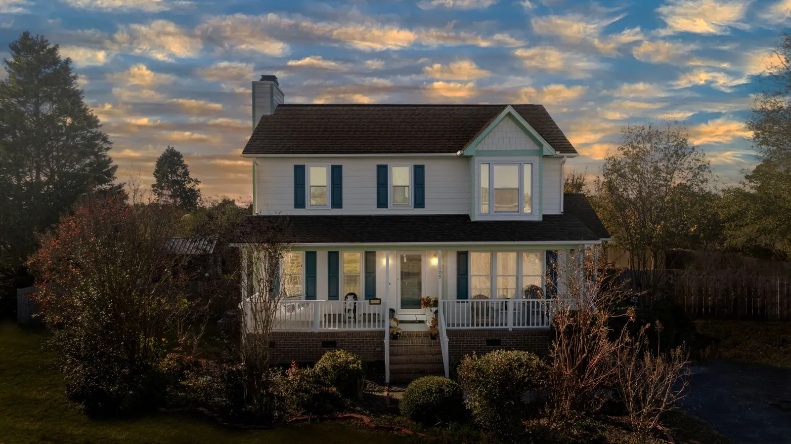 Two-story farmhouse-style home with a covered front porch and glowing windows under a warm sunset sky